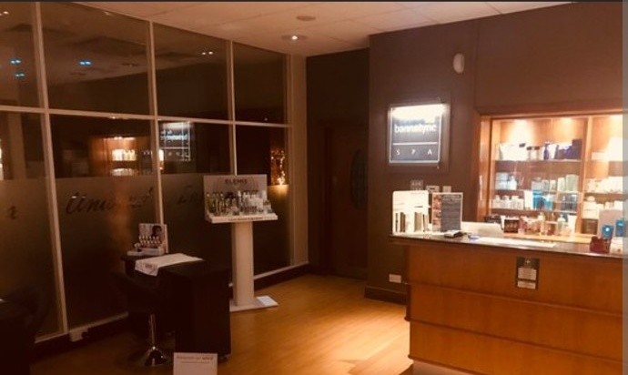 Dimly lit spa reception at Bannatyne Luton, with a wooden counter, illuminated product shelves and a Bannatyne Spa sign
